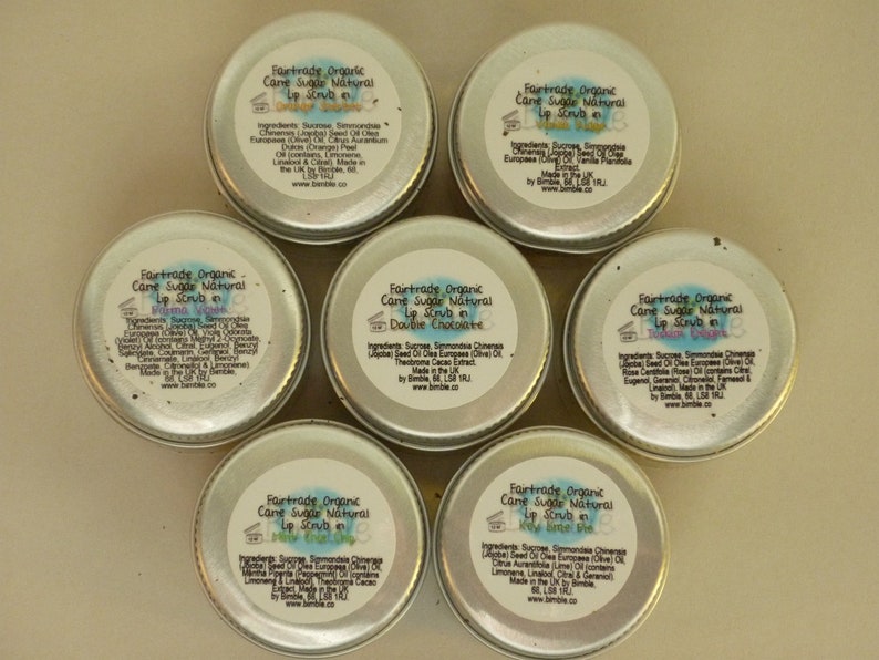 May include: Six small silver tins of Fairtrade Organic Cane Sugar Natural Lip Scrub in various flavours, including Double Chocolate, Prima Vera, and Lemon Sorbet. Each tin has a label with the product name and ingredients.