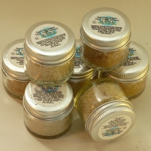 May include: A close-up of seven small glass jars filled with a light brown sugar scrub. The jars have silver lids and white labels with black text that reads "Riverside Organic Cane Sugar Lip Scrub".