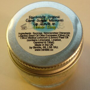 May include: A silver tin with a white label that reads "Fairtrade Organic Cane Sugar Natural Lip Scrub in Lemon Drop". The label also lists the ingredients, which include sucrose, jojoba seed oil, olive oil, lemon peel oil, limonene, linalool, geraniol, and citral. The label also states that the product is made in the UK by Bimble, 68, LS8 1RJ. The label also includes the website address www.bimble.co.