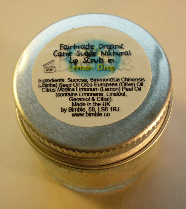 May include: A silver tin with a white label that reads "Fairtrade Organic Cane Sugar Natural Lip Scrub in Lemon Drop". The label also lists the ingredients, which include sucrose, jojoba seed oil, olive oil, lemon peel oil, limonene, linalool, geraniol, and citral. The label also states that the product is made in the UK by Bimble, 68, LS8 1RJ. The label also includes the website address www.bimble.co.