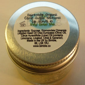 May include: A silver metal jar with a white label that reads "Fairtrade Organic Cane Sugar Natural Lip Scrub in Key Lime Pie". The label also lists the ingredients: Sucrose, Simmondsia Chinensis (Jojoba) Seed Oil Olea Europaea (Olive) Oil, Citrus Aurantifolia (Lime) Oil (contains Limonene, Linalool, Citral & Geraniol). Made in the UK by Bimble, 68, LS8 1RJ. www.bimble.co