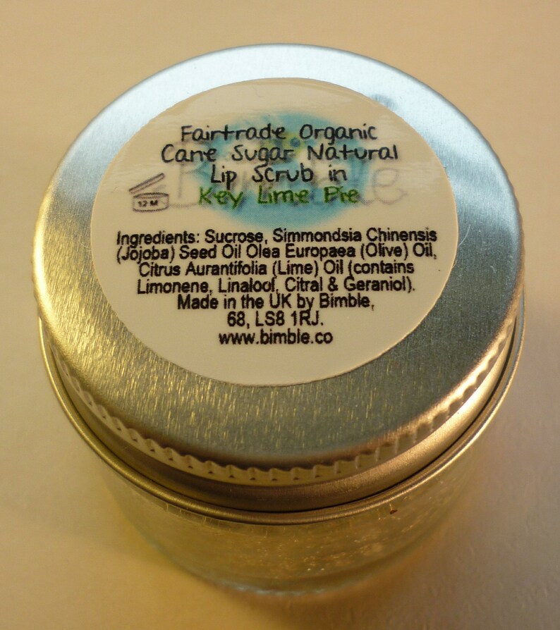 May include: A silver metal jar with a white label that reads "Fairtrade Organic Cane Sugar Natural Lip Scrub in Key Lime Pie". The label also lists the ingredients: Sucrose, Simmondsia Chinensis (Jojoba) Seed Oil Olea Europaea (Olive) Oil, Citrus Aurantifolia (Lime) Oil (contains Limonene, Linalool, Citral & Geraniol). Made in the UK by Bimble, 68, LS8 1RJ. www.bimble.co