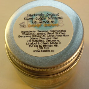 May include: A silver tin with a white label that reads "Fairtrade Organic Cane Sugar Natural Lip Scrub in Orange Sherbet". The label lists the ingredients: Sucrose, Simmondsia Chinensis (Jojoba) Seed Oil Olea Europaea (Olive) Oil, Citrus Aurantium Dulcis (Orange) Peel Oil (contains, Limonene, Linalool & Citral). Made in the UK by Bimble, 68, LS8 1RJ www.bimble.co