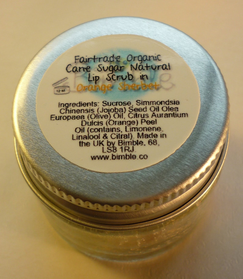 May include: A silver tin with a white label that reads "Fairtrade Organic Cane Sugar Natural Lip Scrub in Orange Sherbet". The label lists the ingredients: Sucrose, Simmondsia Chinensis (Jojoba) Seed Oil Olea Europaea (Olive) Oil, Citrus Aurantium Dulcis (Orange) Peel Oil (contains, Limonene, Linalool & Citral). Made in the UK by Bimble, 68, LS8 1RJ www.bimble.co