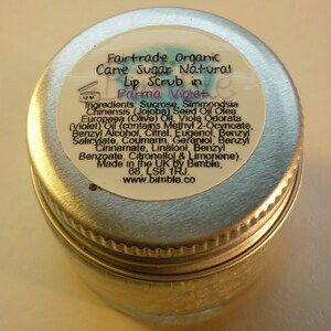 May include: A silver jar with a white label that reads "Fairtrade Organic Cane Sugar Natural Lip Scrub in Parma Violet". The label lists the ingredients, including sucrose, jojoba seed oil, olive oil, and violet oil. The label also states that the product is made in the UK by Bimble.
