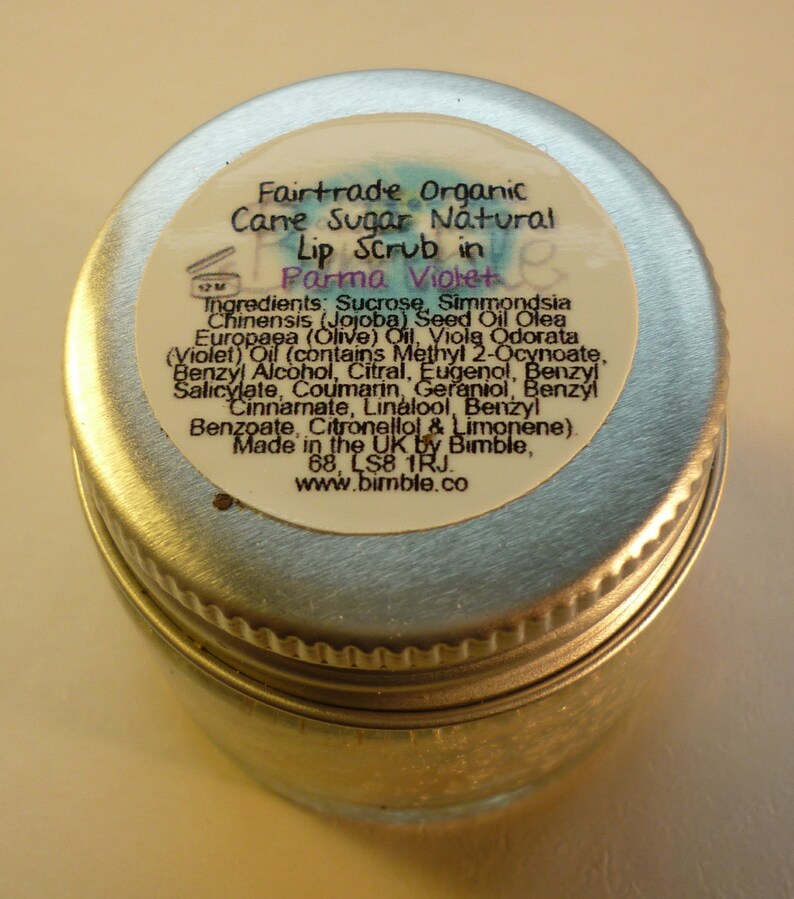 May include: A silver jar with a white label that reads "Fairtrade Organic Cane Sugar Natural Lip Scrub in Parma Violet". The label lists the ingredients, including sucrose, jojoba seed oil, olive oil, and violet oil. The label also states that the product is made in the UK by Bimble.