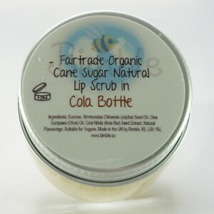 May include: A silver tin with a blue and white label that reads "Fairtrade Organic Cane Sugar Natural Lip Scrub in Cola Bottle". The label also includes a small bee illustration and the text "www.bimble.co".