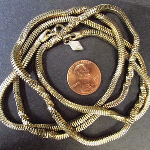 Puede incluir: Un collar de cadena de serpiente en tono dorado con un diseño flexible y segmentado, que incluye una pequeña etiqueta metálica cerca del cierre. Una moneda de un centavo de EE. UU. de color cobre se sitúa en el centro para dar escala.
