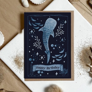 May include: A rectangular greeting card with a dark blue background featuring a white whale shark illustration surrounded by small fish, bubbles, and star shapes. A light blue banner at the bottom contains the text Happy Birthday in dark blue.