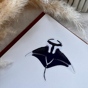 May include: A sticker featuring a black and white illustration of a manta ray with a stippled texture. The manta ray is depicted from a top-down perspective with its wings spread wide and a long, thin tail extending from the centre of its body.