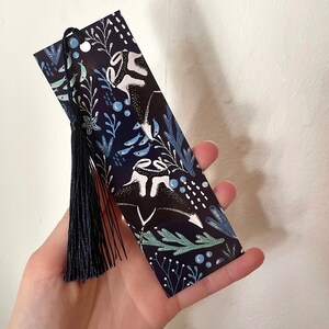 May include: A dark blue bookmark with a white tassel. The bookmark features a whimsical design of stylized white fish and plants on a dark blue background.