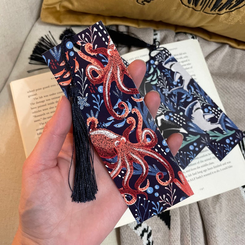 May include: A blue bookmark with a dark blue tassel features a colorful illustration of an octopus with a floral pattern. The octopus is red, orange, and white.
