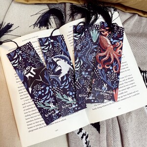 May include: Four black bookmarks with a dark blue background and white and blue illustrations of sea creatures. The bookmarks have a tassel at the top and the text "Kelsey Em" on the bottom. The illustrations include a shark, an octopus, a seahorse, and a manta ray.