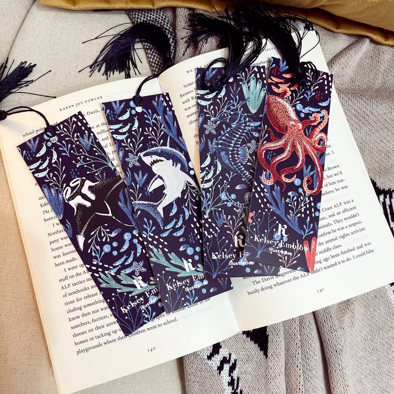 May include: Four black bookmarks with a dark blue background and white and blue illustrations of sea creatures. The bookmarks have a tassel at the top and the text "Kelsey Em" on the bottom. The illustrations include a shark, an octopus, a seahorse, and a manta ray.