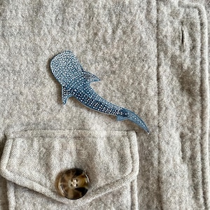 May include: A blue and white whale shark pin is affixed to a gray jacket. The pin is a detailed illustration of the whale shark, with white spots on a blue background. The jacket has a pocket and a button.