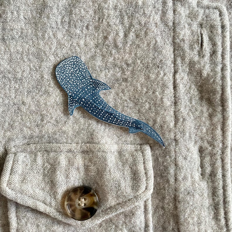 May include: A blue and white whale shark brooch is attached to a grey jacket. The brooch depicts a whale shark with white spots on a blue background. The jacket features a pocket and a button.