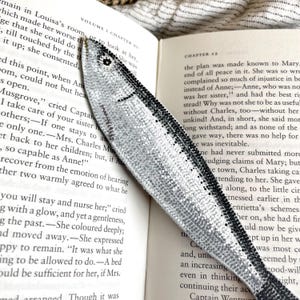 Peut inclure: Un marque-page argent&eacute; en forme de poisson, orn&eacute; de d&eacute;tails noirs, pos&eacute; sur les pages d'un livre. Le marque-page pr&eacute;sente une surface textur&eacute;e et scintillante, ajoutant une touche d&eacute;corative.
