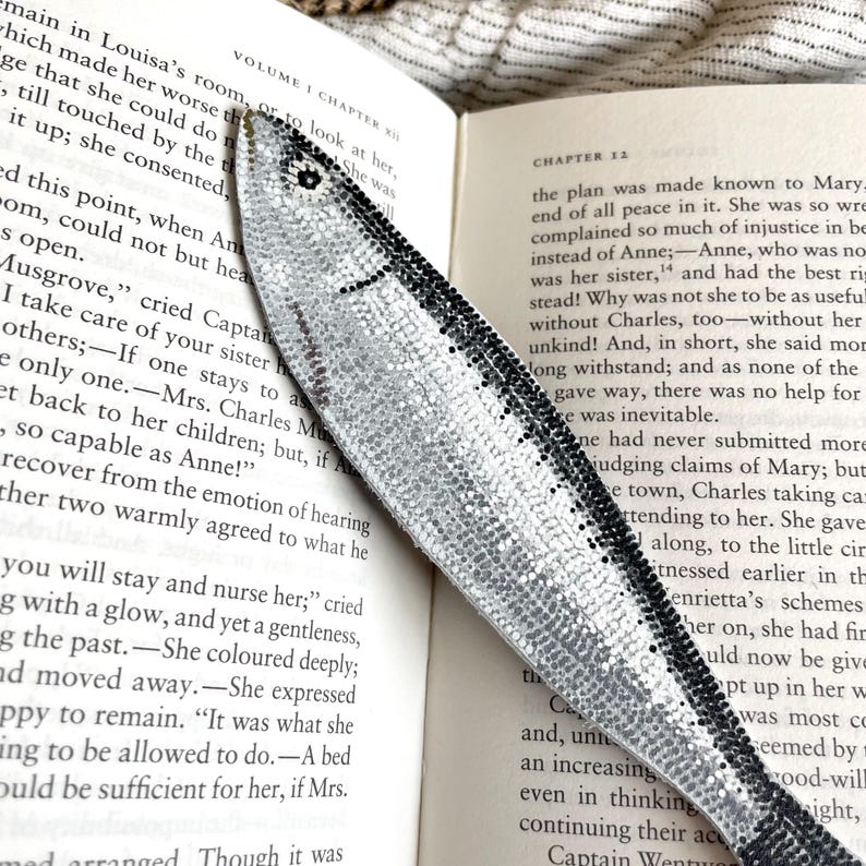 Peut inclure: Un marque-page argent&eacute; en forme de poisson, orn&eacute; de d&eacute;tails noirs, pos&eacute; sur les pages d'un livre. Le marque-page pr&eacute;sente une surface textur&eacute;e et scintillante, ajoutant une touche d&eacute;corative.