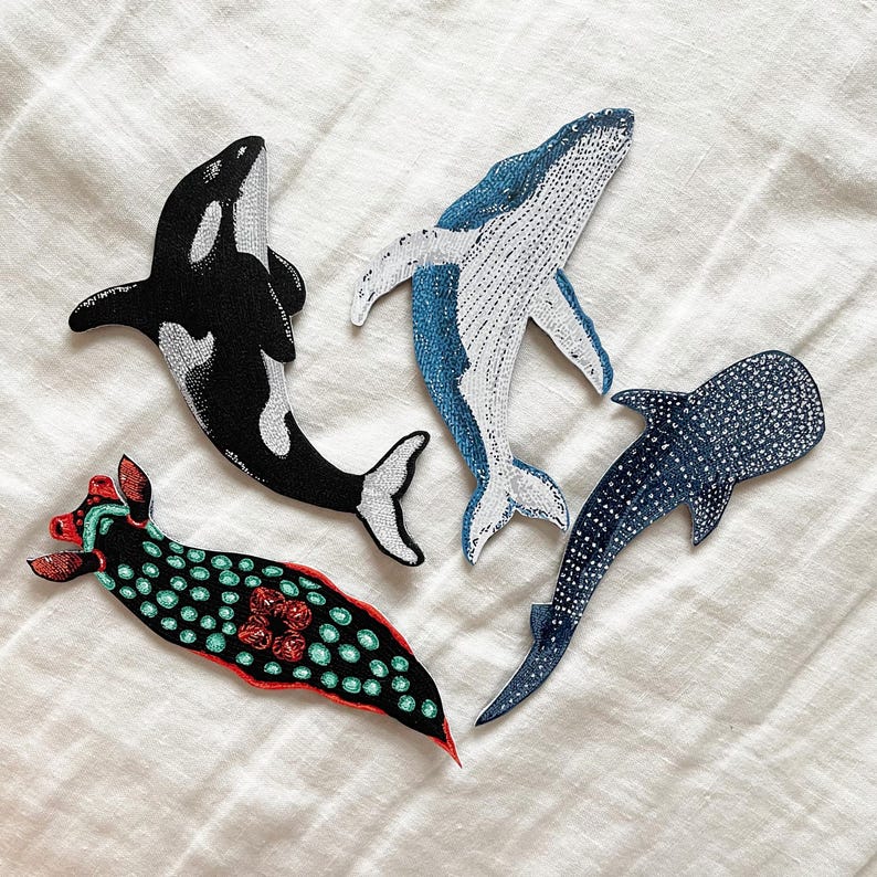 K&ouml;nnte beinhalten: Vier gestickte B&uuml;gelbilder mit Meeresmotiven: ein schwarz-wei&szlig;es Orca, ein blauer Wal, ein blauer und wei&szlig; gefleckter Walhai und eine rot-gr&uuml;ne Nacktschnecke.