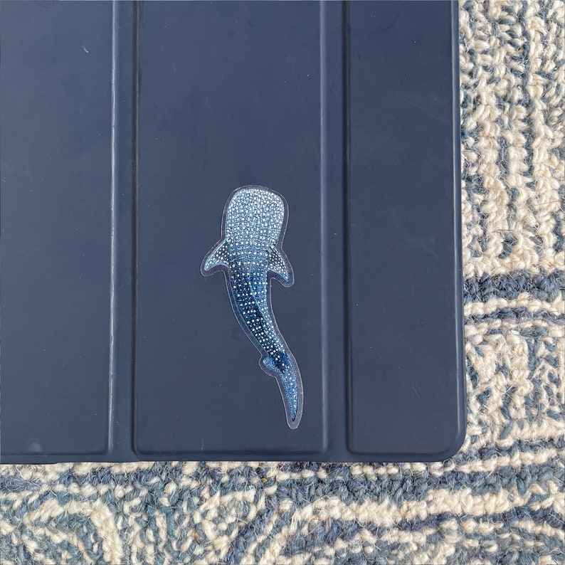 Puede incluir: Una pegatina de tibur&oacute;n ballena azul y blanca sobre un fondo azul oscuro.