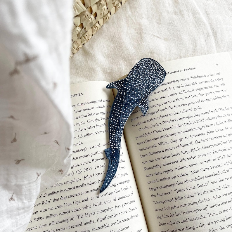 K&ouml;nnte beinhalten: Ein blauer Walhai-Lesezeichen mit silbernen Glitzerakzenten. Das Lesezeichen liegt auf einem Buch mit Text &uuml;ber Social-Media-Marketing und die Macht der sozialen Medien.
