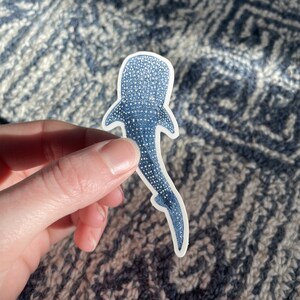 Puede incluir: Una pegatina de tibur&oacute;n ballena azul con un contorno blanco. La pegatina est&aacute; sobre una tela con estampado azul y blanco.