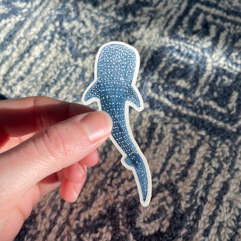 Puede incluir: Una pegatina de tibur&oacute;n ballena azul con un contorno blanco. La pegatina est&aacute; sobre una tela con estampado azul y blanco.