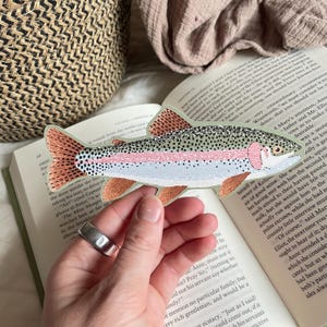 Op de afbeelding: Geborduurde patch in de vorm van een regenboogforel. De vis heeft een witte buik, een roze streep en een gespikkelde groene rug. De staart en vinnen zijn oranje. De patch wordt boven een open boek gehouden.