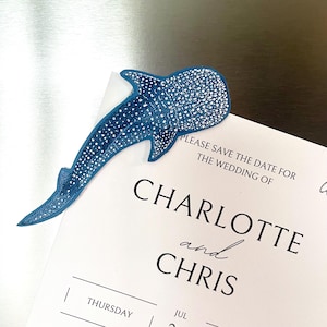 May include: A blue whale shark magnet with a white spotted pattern placed on a white save the date card that reads: PLEASE SAVE THE DATE FOR THE WEDDING OF CHARLOTTE and CHRIS THURSDAY JUL.