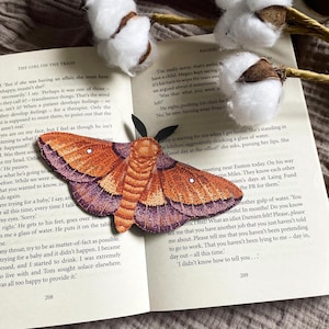 Puede incluir: Una detallada mariposa naranja y morada de papel recortado descansa sobre un libro abierto. La mariposa tiene intrincados patrones en las alas y está cerca de bolas de algodón. Las páginas del libro muestran texto impreso.
