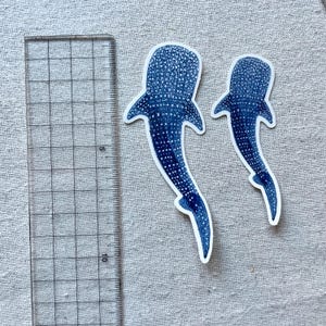 Puede incluir: Dos pegatinas de tibur&oacute;n ballena azul sobre un fondo de tela blanca. Las pegatinas miden aproximadamente 15 cm y 10 cm de largo.