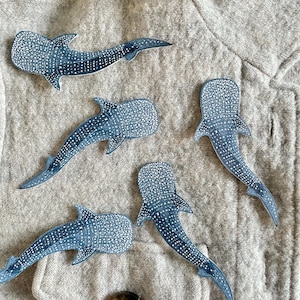 May include: Five blue whale shark cutouts with white dot patterns are arranged on a textured, light grey surface. The whale sharks have detailed fins and tails, and are in various positions.