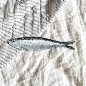 Peut inclure: Un patch de sardine brod&eacute;e argent et noir. Le poisson pr&eacute;sente un motif d'&eacute;cailles d&eacute;taill&eacute; et une bande dorsale noire. Le patch est pos&eacute; sur un fond de tissu blanc et ray&eacute;.