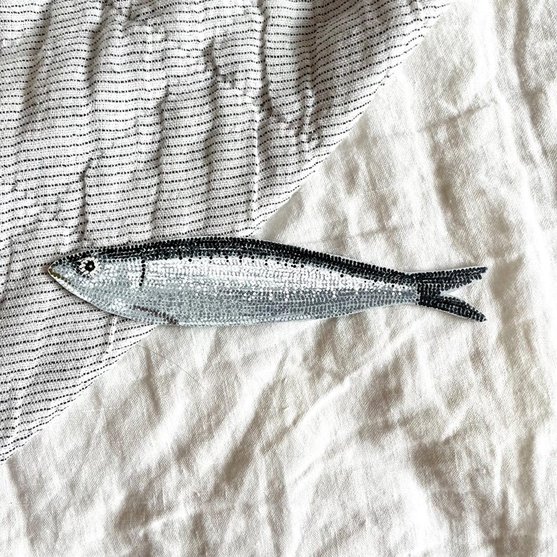 Peut inclure: Un patch de sardine brod&eacute;e argent et noir. Le poisson pr&eacute;sente un motif d'&eacute;cailles d&eacute;taill&eacute; et une bande dorsale noire. Le patch est pos&eacute; sur un fond de tissu blanc et ray&eacute;.