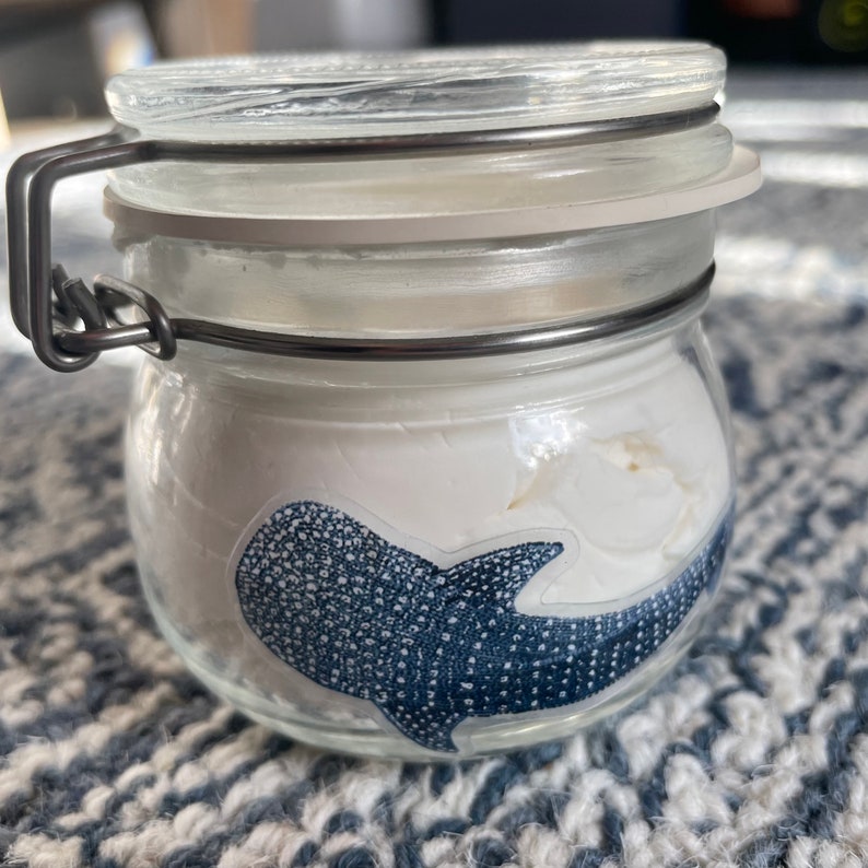 Puede incluir: Un tarro de vidrio transparente con tapa de metal y una pegatina de tibur&oacute;n ballena azul y blanca en la parte delantera.