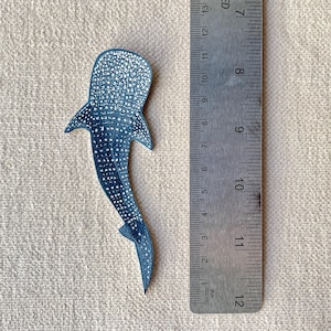 May include: A dark blue whale shark-shaped item with white spots, next to a metal ruler. The item is approximately 12 cm long. The ruler shows metric and imperial measurements. The background is a light tan fabric.
