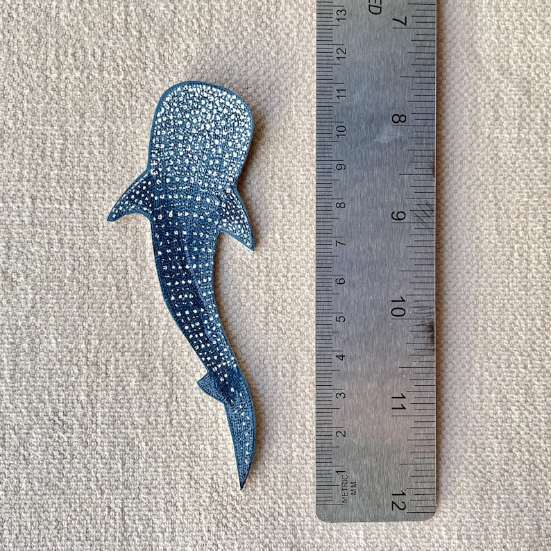 May include: A dark blue whale shark-shaped item with white spots, next to a metal ruler. The item is approximately 12 cm long. The ruler shows metric and imperial measurements. The background is a light tan fabric.