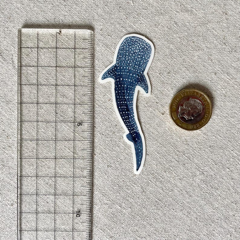 Puede incluir: Una pegatina de tibur&oacute;n ballena azul y blanca con un patr&oacute;n de manchas. La pegatina mide aproximadamente 5 cm de largo.