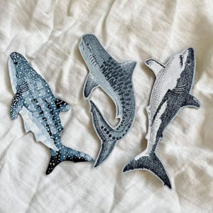 Peut inclure: Trois &eacute;cussons en forme de requin dans des tons de bleu et de blanc. Les &eacute;cussons pr&eacute;sentent des illustrations d&eacute;taill&eacute;es de diff&eacute;rentes esp&egrave;ces de requins, avec des motifs et des textures vari&eacute;s. Les &eacute;cussons sont plac&eacute;s sur une surface blanche et textur&eacute;e.
