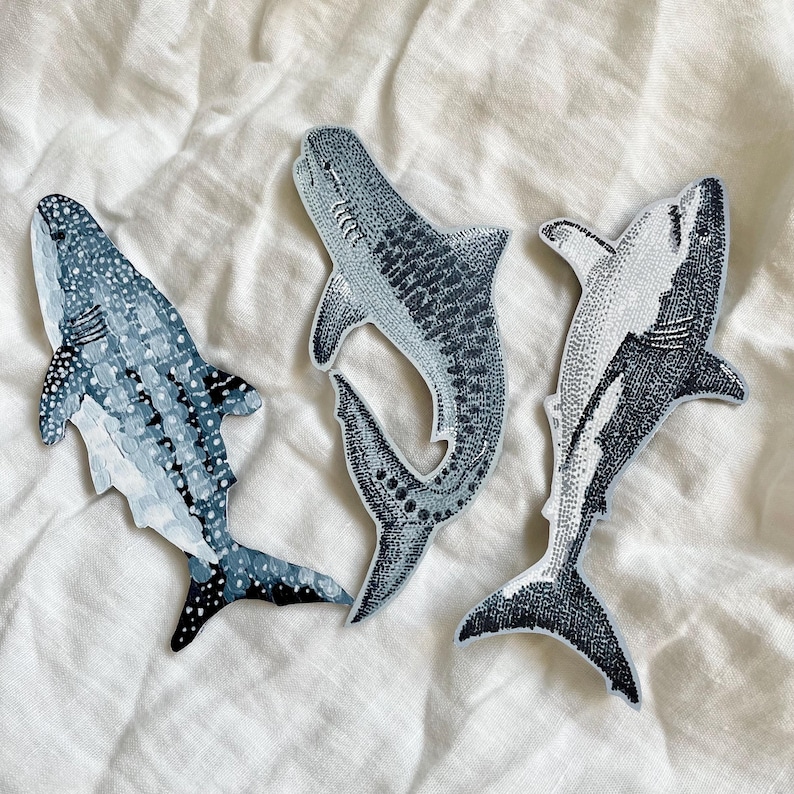 Peut inclure: Trois &eacute;cussons en forme de requin dans des tons de bleu et de blanc. Les &eacute;cussons pr&eacute;sentent des illustrations d&eacute;taill&eacute;es de diff&eacute;rentes esp&egrave;ces de requins, avec des motifs et des textures vari&eacute;s. Les &eacute;cussons sont plac&eacute;s sur une surface blanche et textur&eacute;e.