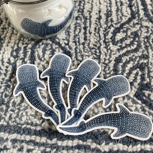 Puede incluir: Cinco pegatinas de tibur&oacute;n ballena azul y blanco sobre un fondo de tela a rayas blanco y gris. Las pegatinas est&aacute;n dispuestas en semic&iacute;rculo con las colas apuntando hacia el centro.