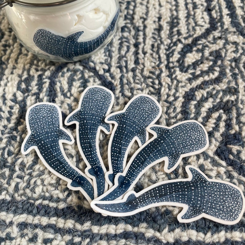 Puede incluir: Cinco pegatinas de tibur&oacute;n ballena azul y blanco sobre un fondo de tela a rayas blanco y gris. Las pegatinas est&aacute;n dispuestas en semic&iacute;rculo con las colas apuntando hacia el centro.
