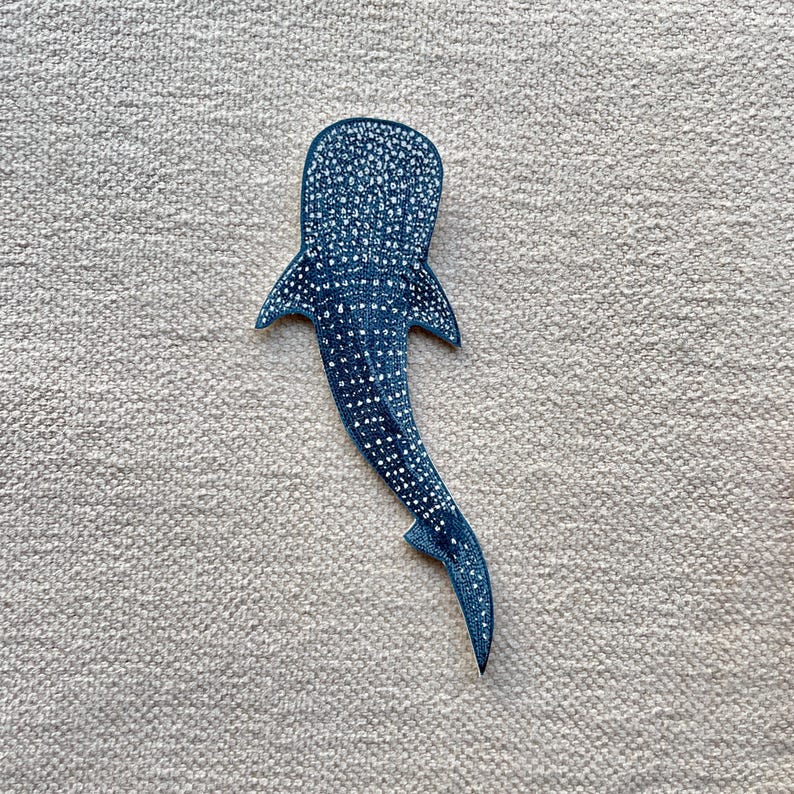 May include: A blue whale shark patch with white spots, mimicking the pattern of a whale shark. The fabric patch features a detailed design. The patch is set on a textured, light grey surface.
