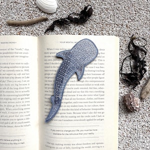 May include: A blue whale shark patch with a white spotted pattern and a textured surface, positioned vertically on the open pages of a book. The patch features a detailed silhouette of the shark with visible fins and a tapered tail section.