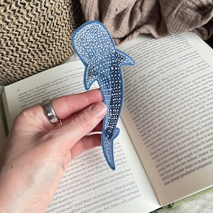 Puede incluir: Un parche de tiburón ballena azul con un patrón detallado de puntos blancos y un borde azul oscuro. El accesorio textil bordado tiene la forma de la criatura marina, mostrando una cabeza ancha, aletas laterales y una cola larga y estrecha.
