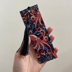May include: A black bookmark with a tassel featuring a colorful illustration of two octopuses with blue, red, and white accents. The bookmark has a dark blue background with white and light blue details.