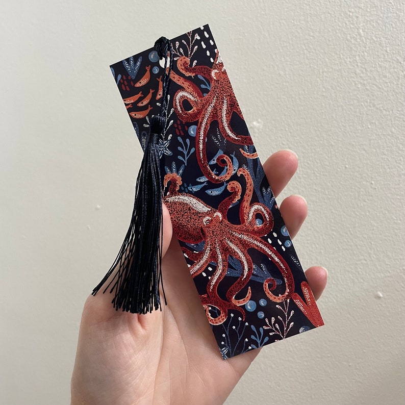 May include: A black bookmark with a tassel featuring a colorful illustration of two octopuses with blue, red, and white accents. The bookmark has a dark blue background with white and light blue details.