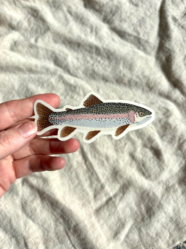 May include: A colorful sticker of a rainbow trout. The fish has a gray body with a pink stripe, black spots, and brown fins. The sticker is on a white background.