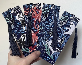 May include: Set of four blue and white bookmarks with colourful illustrations of sea creatures and tassels. The bookmarks feature an octopus, a fish, a seahorse, and a shark.