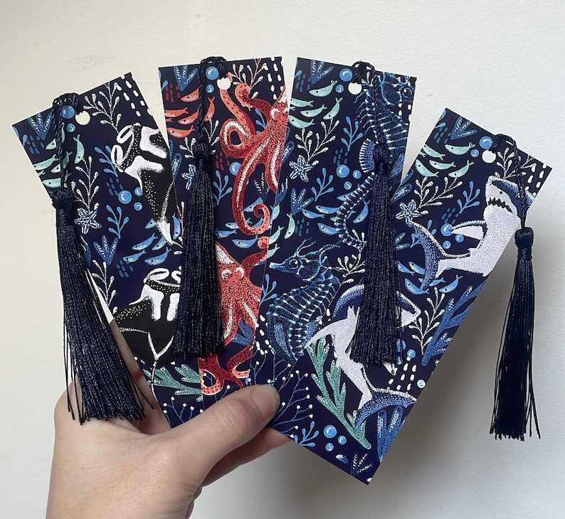 May include: Set of four blue and white bookmark with colorful illustrations of sea creatures and tassels. The bookmarks feature an octopus, a fish, a seahorse, and a shark.